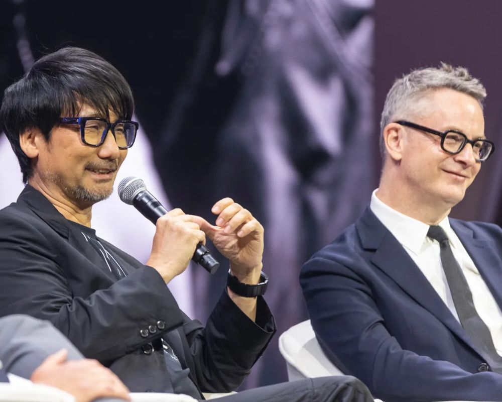 Hideo Kojima hablando en el escenario de la conferencia New Global Sport 2025, con el logotipo de Kojima Productions en la pantalla de fondo