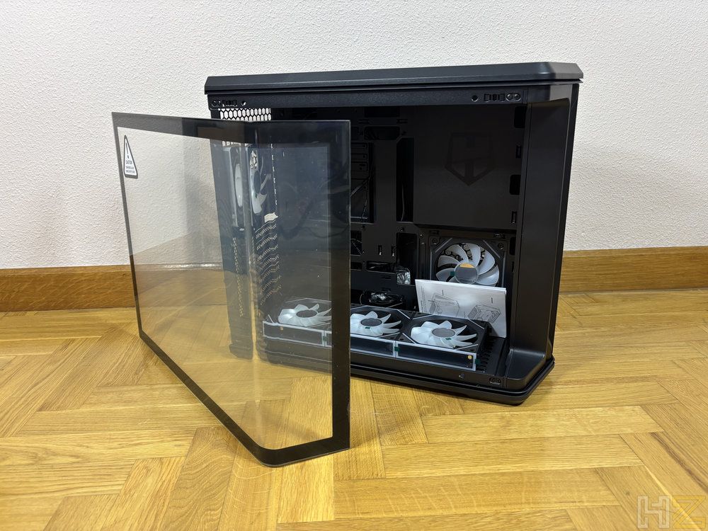 Caja de ordenador Nox Hummer Anvil abierta sobre suelo de parqué, mostrando el panel lateral de cristal templado desmontado y ventiladores internos.