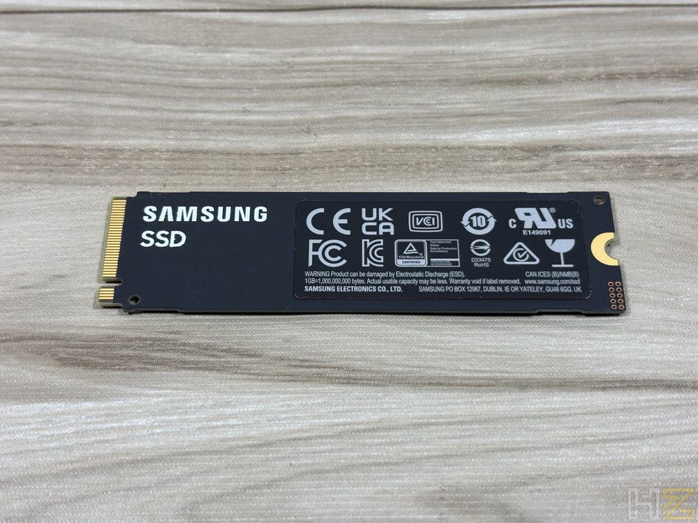 Unidad de almacenamiento SSD Samsung NVMe sobre una superficie de madera clara, mostrando su etiqueta negra con certificaciones y conectores dorados.