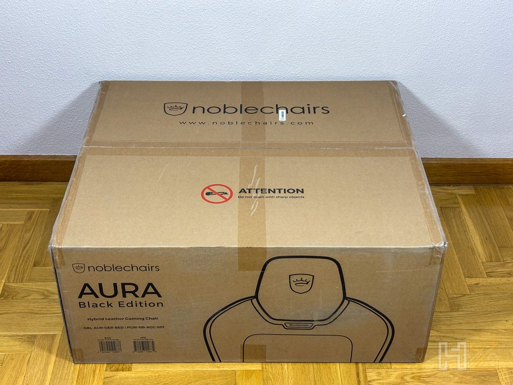 Caja de embalaje de cartón marrón de la silla gaming noblechairs AURA Black Edition apoyada sobre un suelo de parquet.