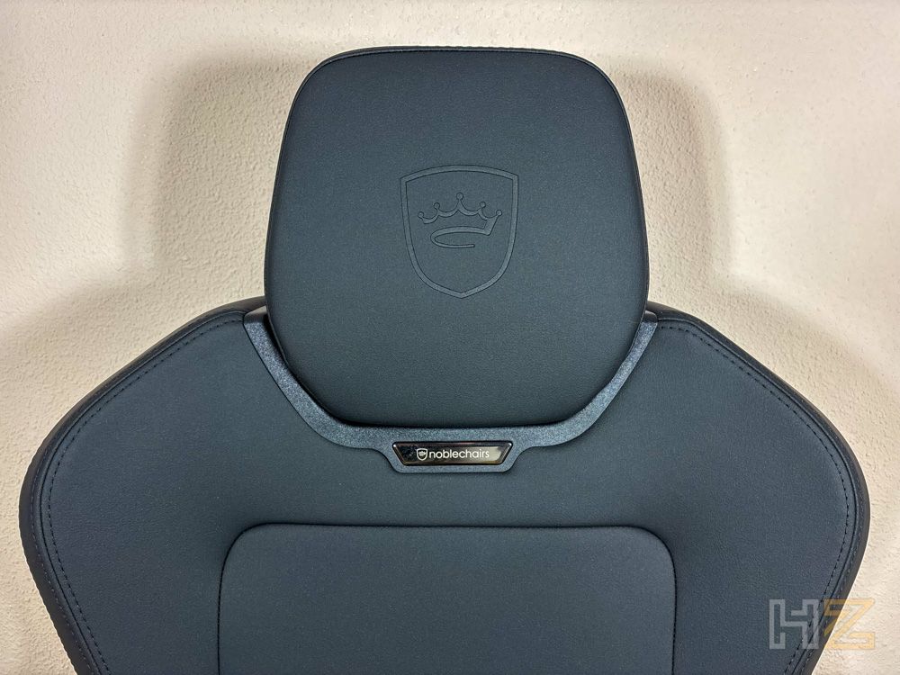 Primer plano del reposacabezas de una silla noblechairs AURA en color negro con el logotipo de la corona grabado en la piel y una placa metálica distintiva en la base del apoyo.